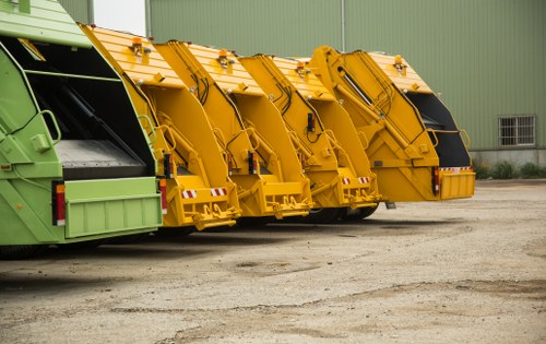 A row of four large waste collection vehicles parked on a gravel surface in front of a green industrial building. The first vehicle on the left is painted in a pale green colour, while the remaining three are bright yellow. Each vehicle is equipped with a large, enclosed hopper with a hydraulic lifting mechanism, designed for collecting and disposing of waste. The vehicles are positioned side by side, with the green one on the far left, and the yellow ones lined up next, facing towards the right. The gravel surface is level, with no visible debris, and the building behind has metal siding with a small window. The scene appears to be part of a commercial waste collection yard, possibly servicing local businesses in the Purfleet area, near the postcode district inferred from the 'Commercial Waste Purfleet' service page, illustrating the fleet used by Commercial Waste Purfleet in the rubbish removal industry.