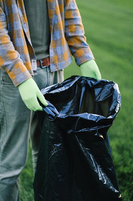 On a paved sidewalk area beside a black metal fence with vertical bars and leafy trees in the background, three large black plastic rubbish bags filled with waste are stacked against the fence. The ba